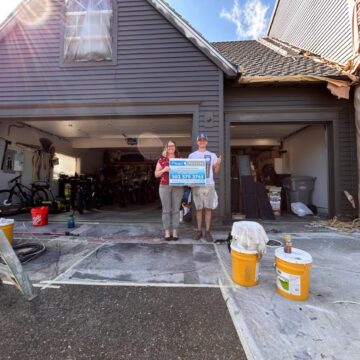 Renee & Seamus Barron hold a Pearl Painters sign by a garage under repair in Portland, with tools and materials around.