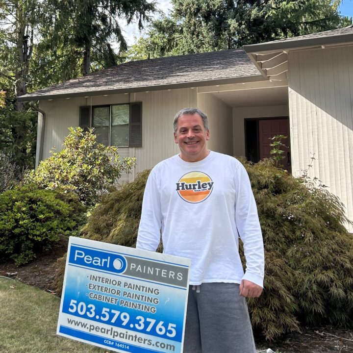 Man in white Hurley shirt holds Pearl Painters sign with contact info outside beige house, likely a Portland painting company.