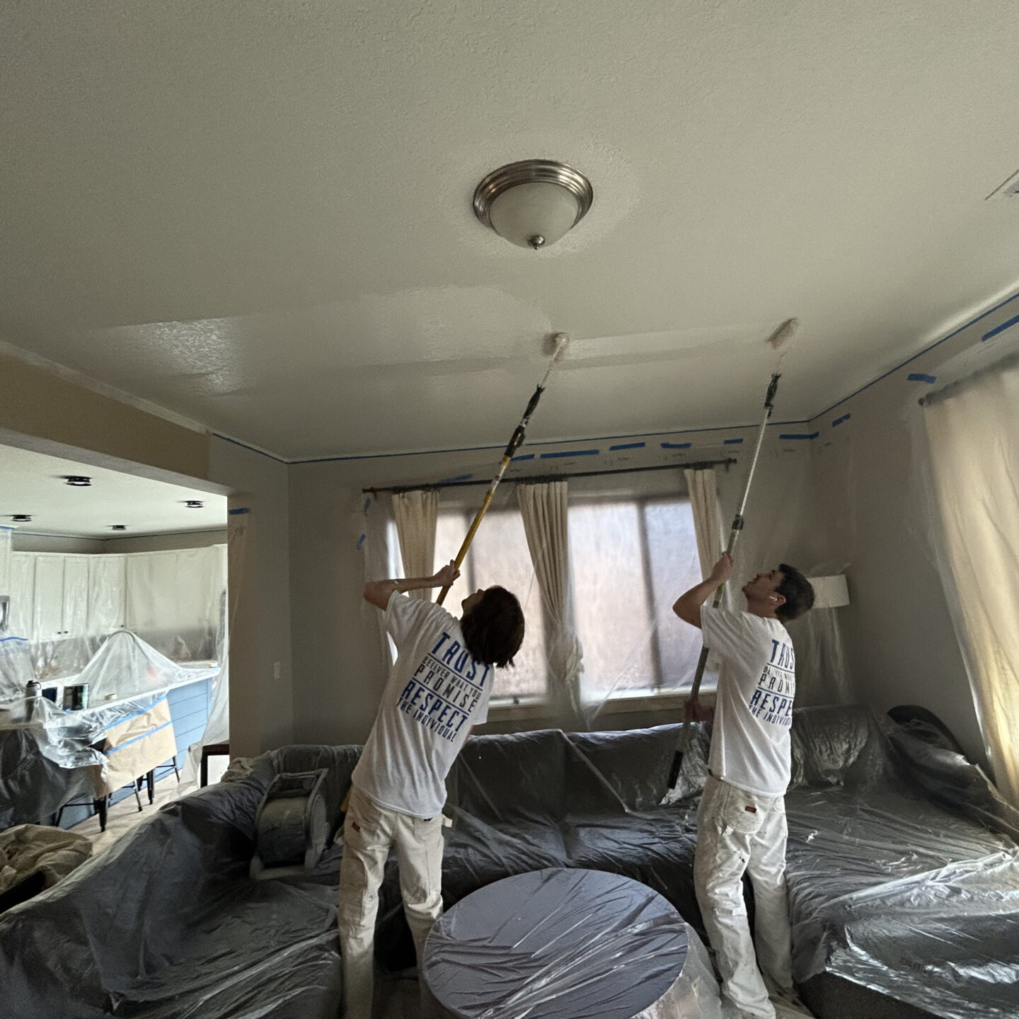 Painters use rollers on interior ceilings in Portland as Pearl Painters; furniture and windows covered with plastic and tape.