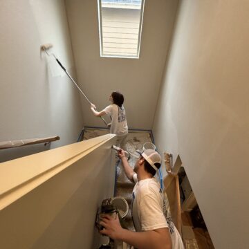 Two painters in white shirts use rollers on a stairwell; a skylight lights the Portland interior painting project by Pearl Painters.