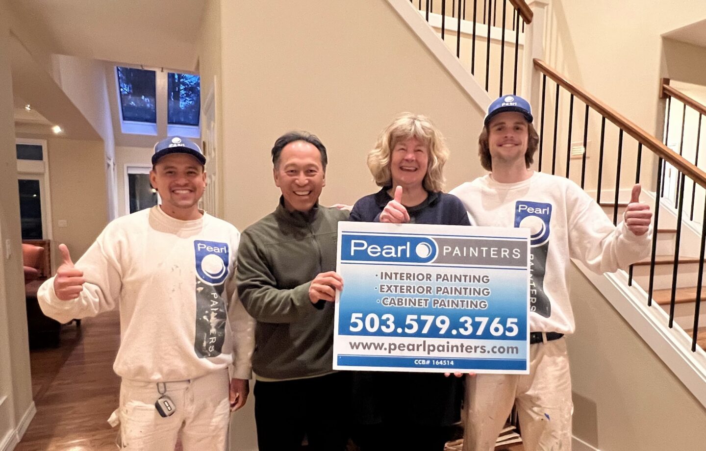 Two painters and a couple give thumbs up while holding a Pearl Painters sign in a well-lit home entryway. The sign includes services: interior, exterior, and cabinet painting with a contact number.