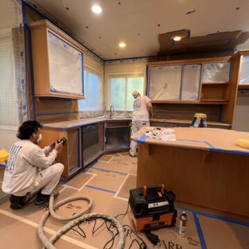 Two painters in white overalls working in a kitchen covered with protective plastic sheets and tape. one is painting a cabinet while the other prepares equipment.