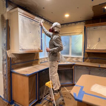 A construction worker in protective gear paints a wall inside a room covered with protective plastic sheeting and blue painter's tape.