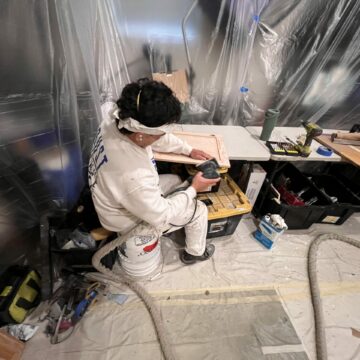 A worker in a protective suit sands a wooden board in a room covered with plastic sheeting.