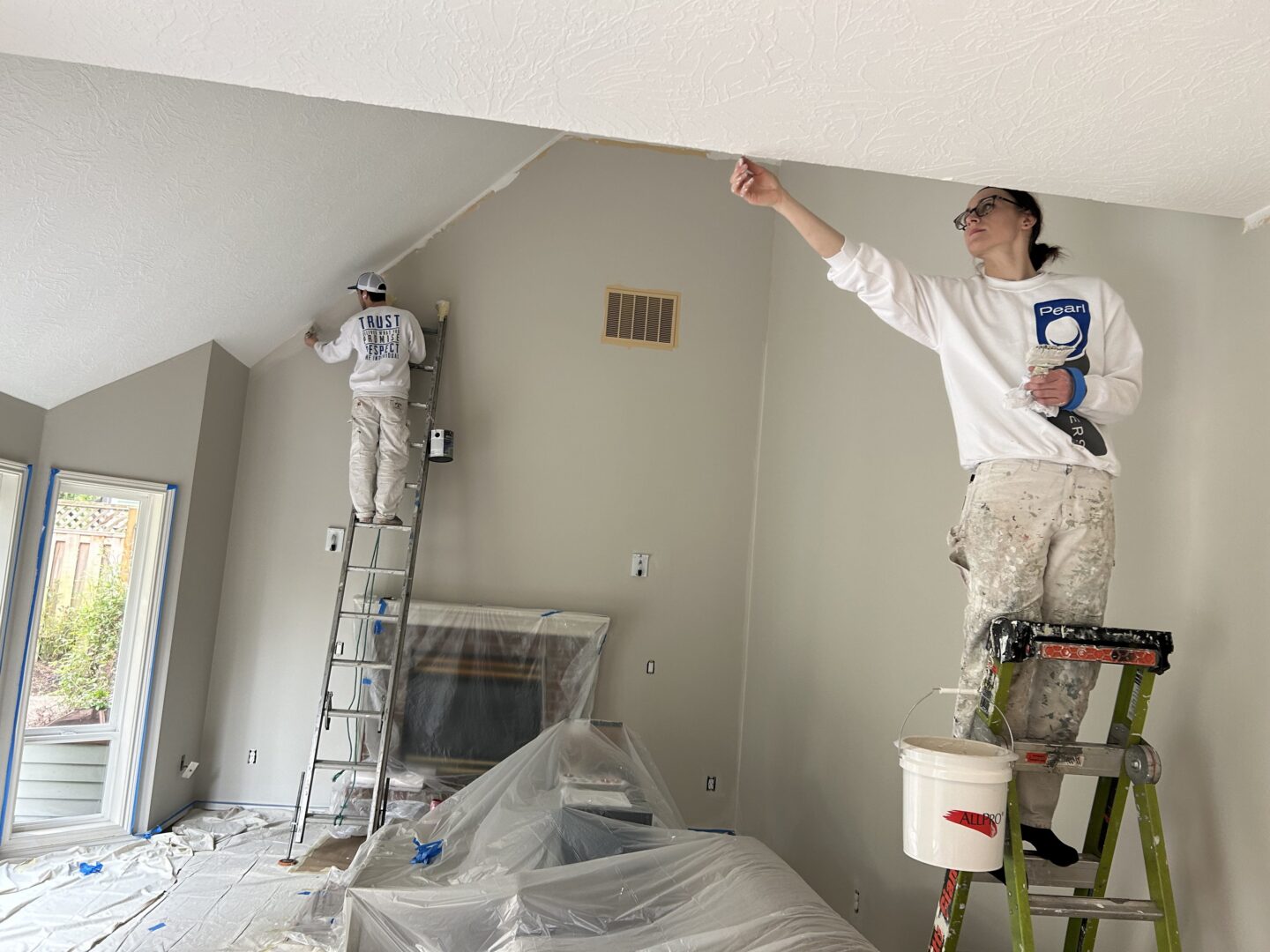 Painters on ladders paint a living room; Pearl Painters cover the floor and furniture with sheets for a tidy job in Portland.