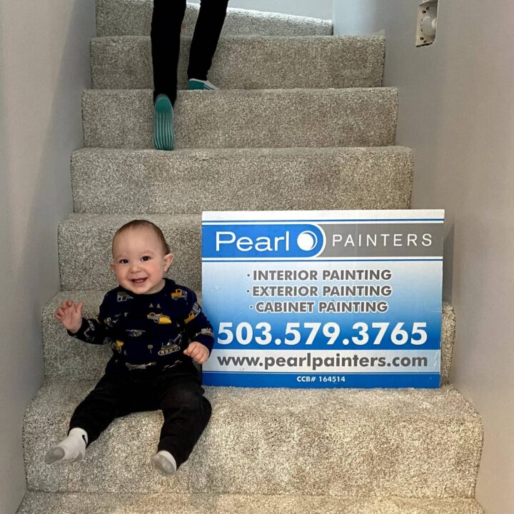 Baby on carpeted stairs beside a Pearl Painters sign with contact info; adult feet above. Painting company is in Portland.