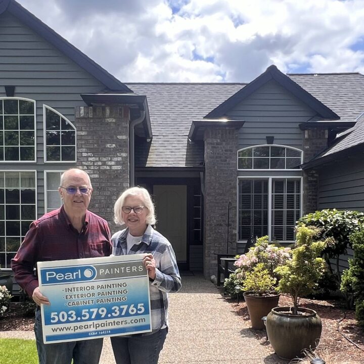 Two people hold a Pearl Painters sign in front of a house in Portland, advertising painting services with contact info.