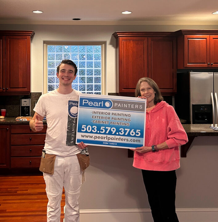 A painter and woman in a kitchen in Portland hold a Pearl Painters sign with contact info and painting service details.