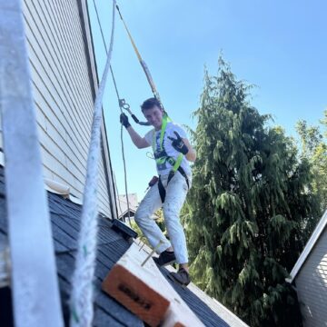 A painter in safety gear stands on a sloped roof in Portland, secured by harness; painting by Pearl Painters, August 2025.