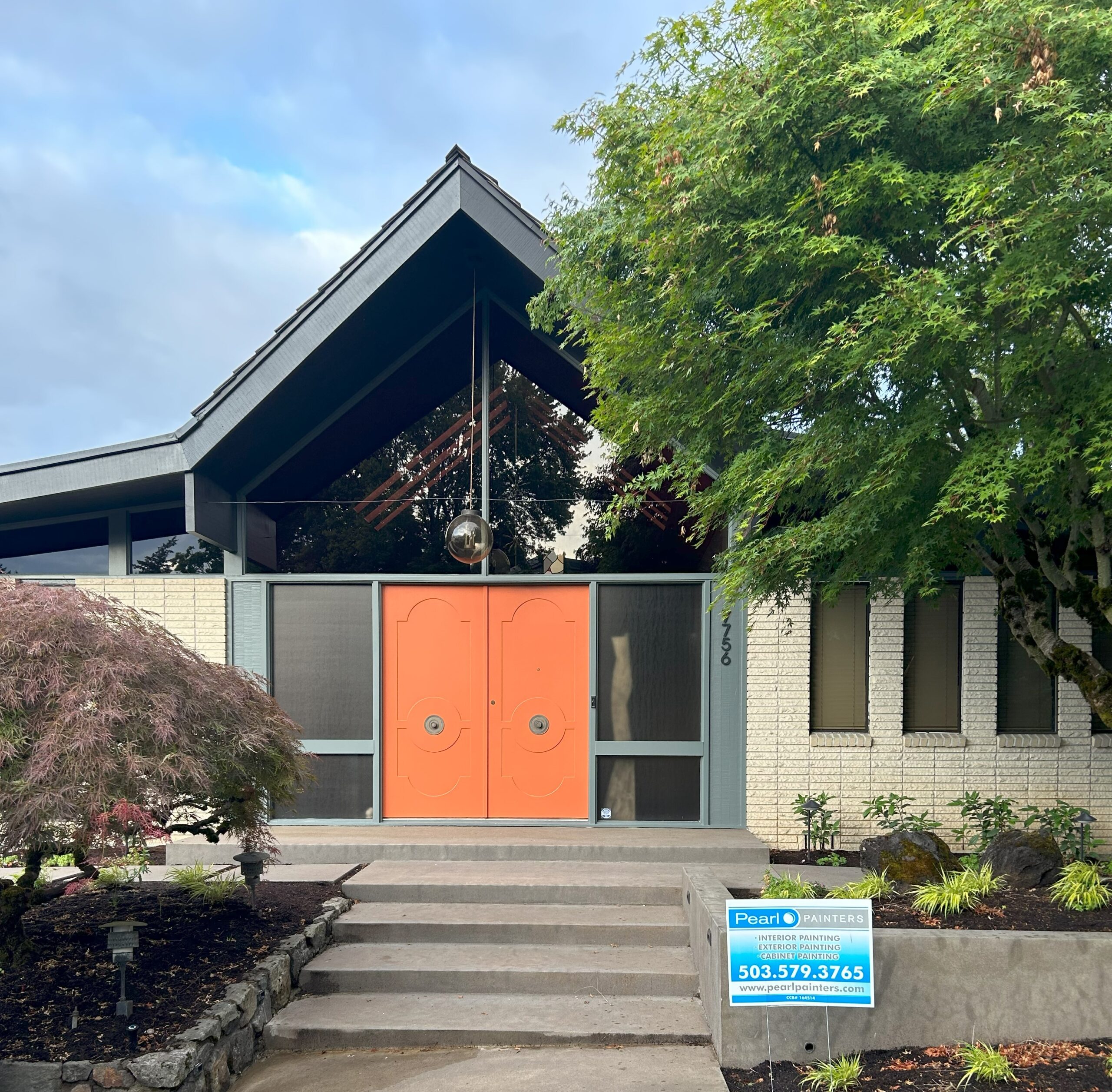 A modern house with a steeply pitched roof, orange double doors, and a lush green tree in the front yard invites you in. A sign near the entrance displays a painting company’s contact information, reminding passersby to book early for their next exterior project before the heat sets in.