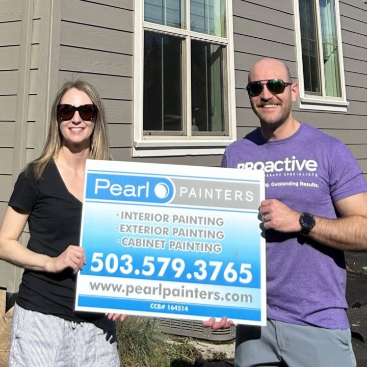 Two people outside in sunlight hold a Pearl Painters sign for painting services in Portland, including contact info.