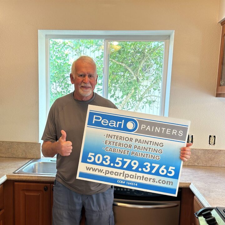 Man in kitchen holds Pearl Painters sign for painting services—interior, exterior, cabinets—in Portland with contact info.