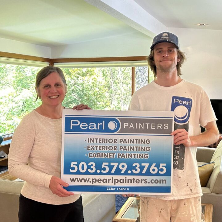 Two people indoors hold a Pearl Painters sign for painting services in Portland, showing phone number and website.