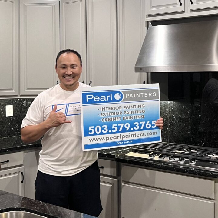 Person in kitchen holds Pearl Painters sign for interior, exterior, and cabinet painting in Portland; phone and website shown.