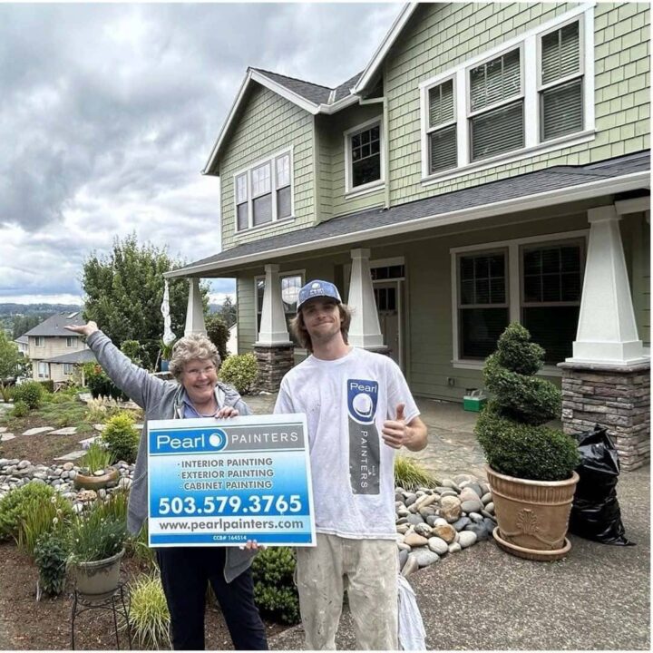 Two people hold a Pearl Painters painting sign in front of a green house with shrubs and rocks, likely in Portland.