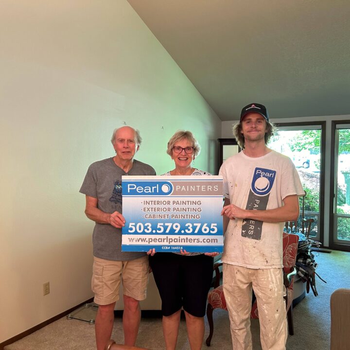 Three people indoors in Portland hold a Pearl Painters sign; one wears painter's overalls. Light walls and large windows behind them.