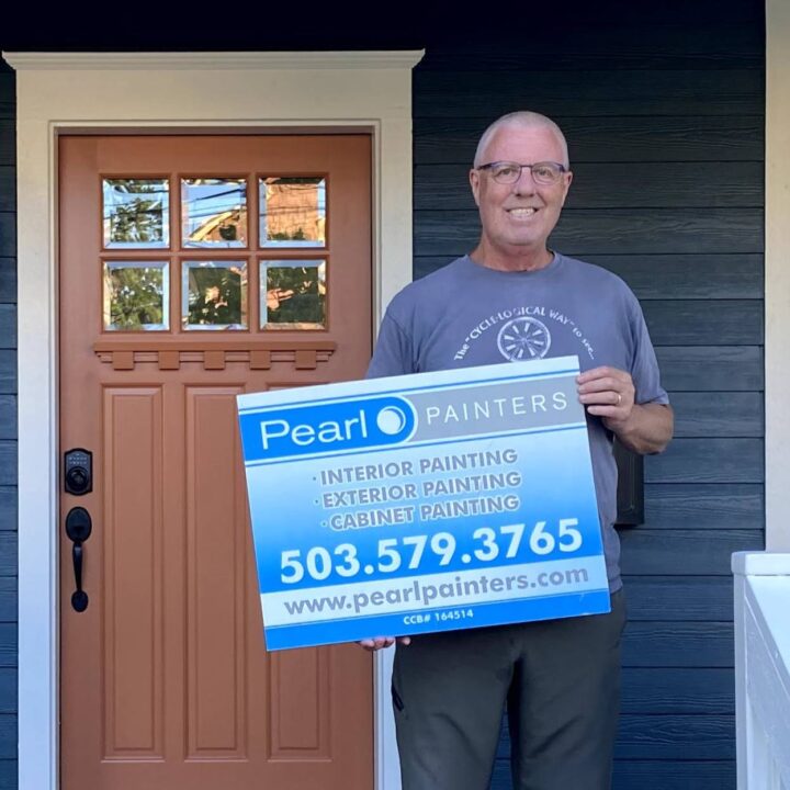 Man in front of blue house in Portland holds Pearl Painters sign for painting services with contact info.