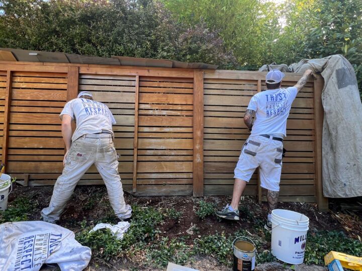 Two people are busy with fence & deck staining, dressed in white clothes and hats. Paint buckets are placed nearby, while one person partially covers the fence with a tarp. Trees provide a serene backdrop to their industrious task.