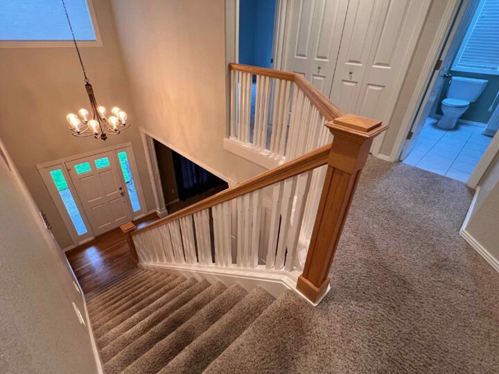 From the top of carpeted stairs with a sleek Bull Mountain Handrail, gaze down to a market-ready foyer featuring a stylish chandelier and front door. A bathroom with a toilet is visible on the right, enhancing the modern look of this inviting space.