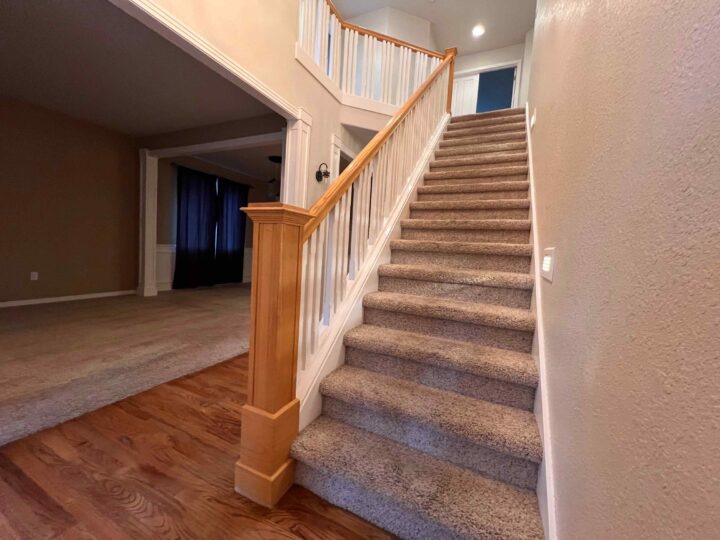 A carpeted staircase with a Bull Mountain Handrail leads to the second floor of a house. The main floor boasts both hardwood and carpeted flooring, complemented by an open doorway to another room, showcasing a market-ready design.