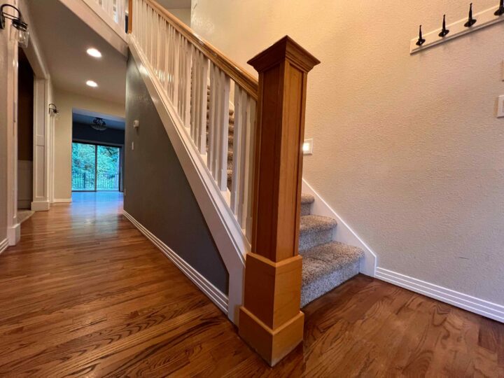 The hallway boasts a modern look with sleek wooden floors and a staircase featuring carpeted steps. A view of the lit room showcases large glass doors leading outside, complemented by the elegant Bull Mountain Handrail for a market-ready appeal.