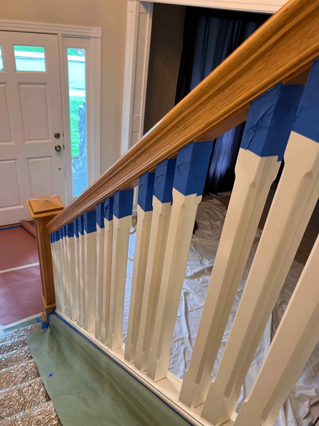 A wooden staircase with white balusters and a Bull Mountain handrail is partially covered in blue painter's tape, indicating ongoing painting work. Protective sheets cover the floor, promising a market-ready, modern look upon completion.