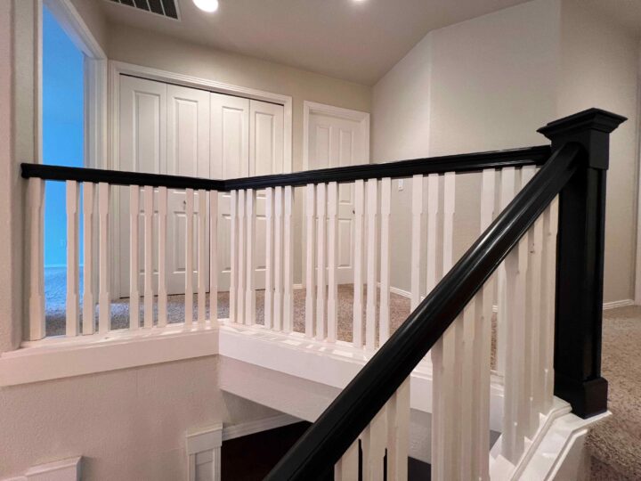 The staircase features white balusters and a Bull Mountain Handrail with a dark finish, leading to a hallway of closed doors. A carpeted floor and neutral walls complete the scene.