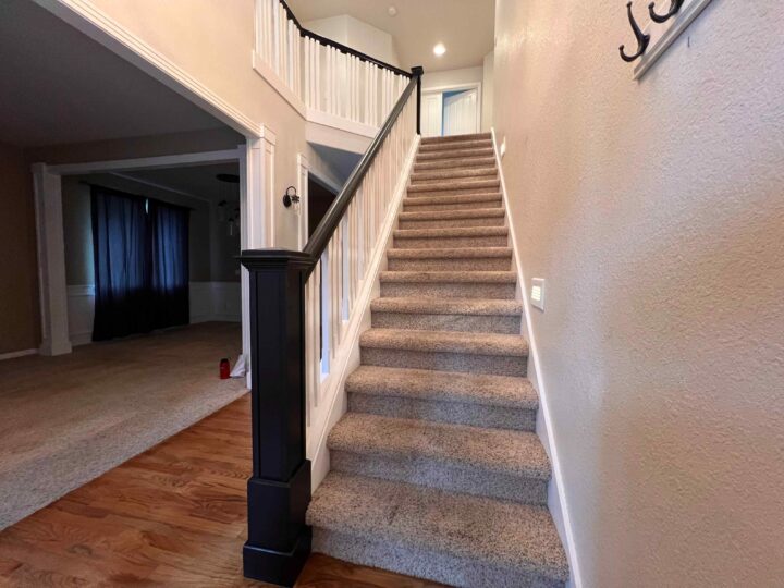 A carpeted staircase with a Bull Mountain Handrail in black and white leads to an upper floor in a house. The hallway, boasting beige walls, connects to a market-ready carpeted room with dark curtains.