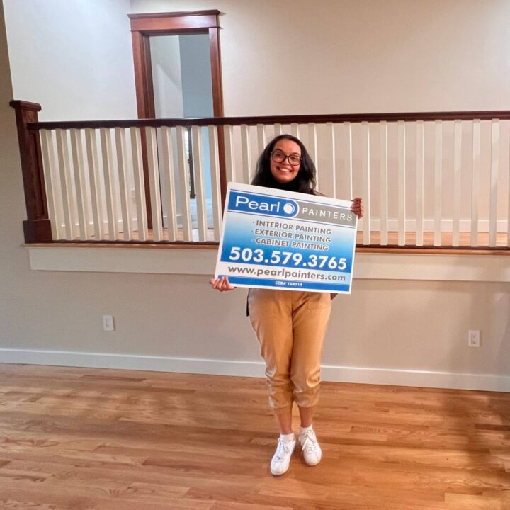 Woman in a room with wooden floors holds a Pearl Painters sign for painting services in Portland, showing phone number and website.