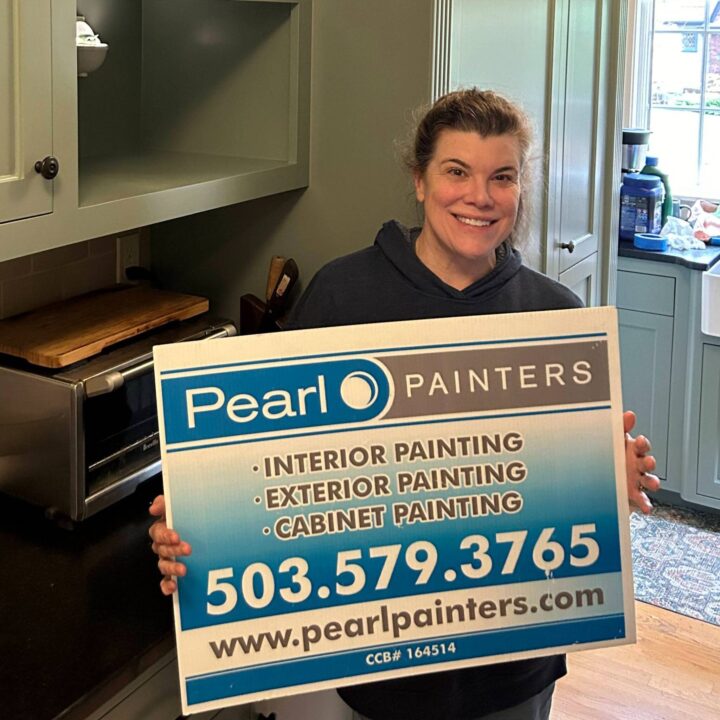 Woman in a kitchen holds a Pearl Painters sign for interior, exterior, and cabinet painting in Portland with contact info.