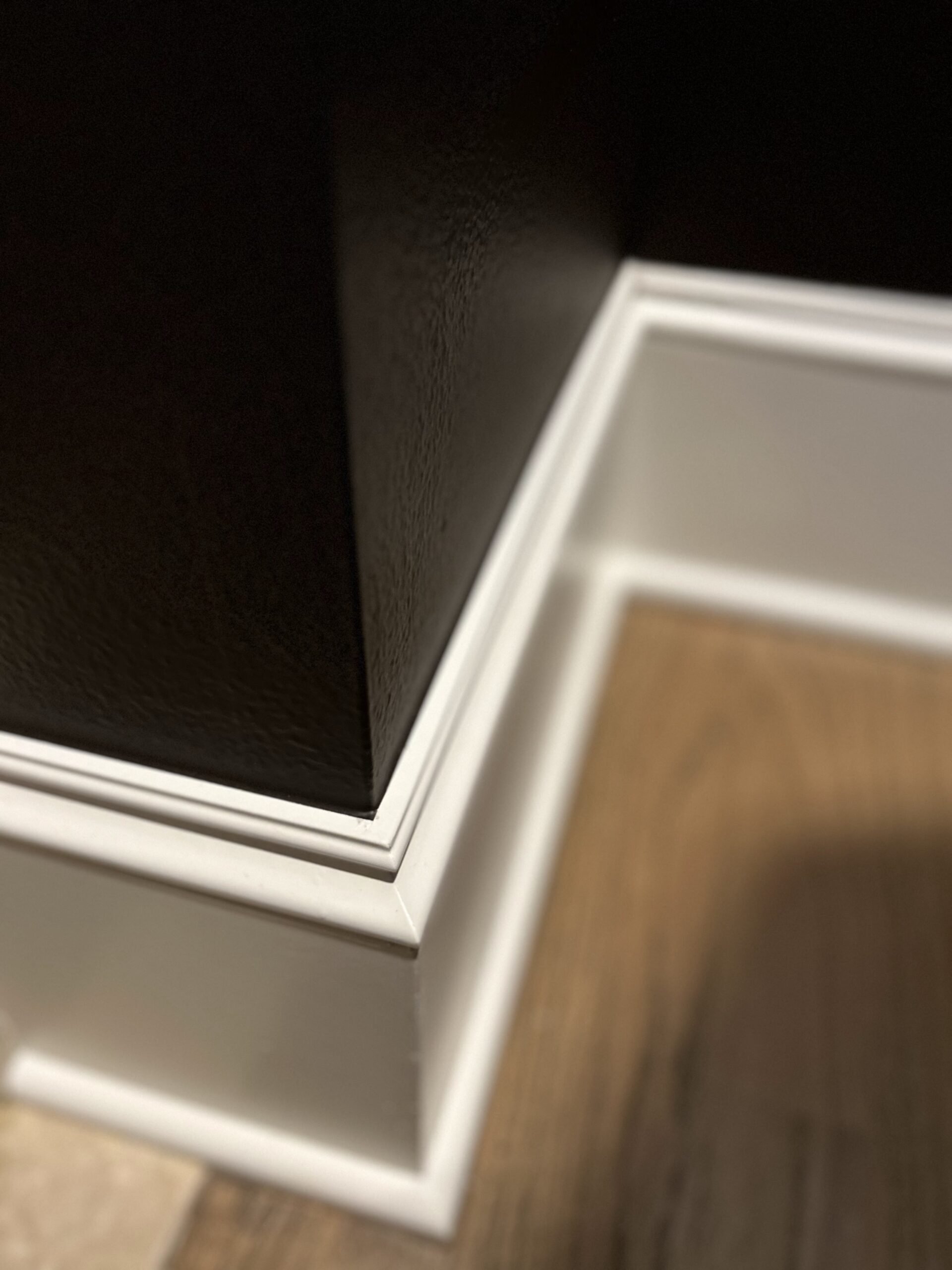 White baseboard trim glows against the black wall and wood floor corner, showcasing a properly painted surface by Pearl Painters.