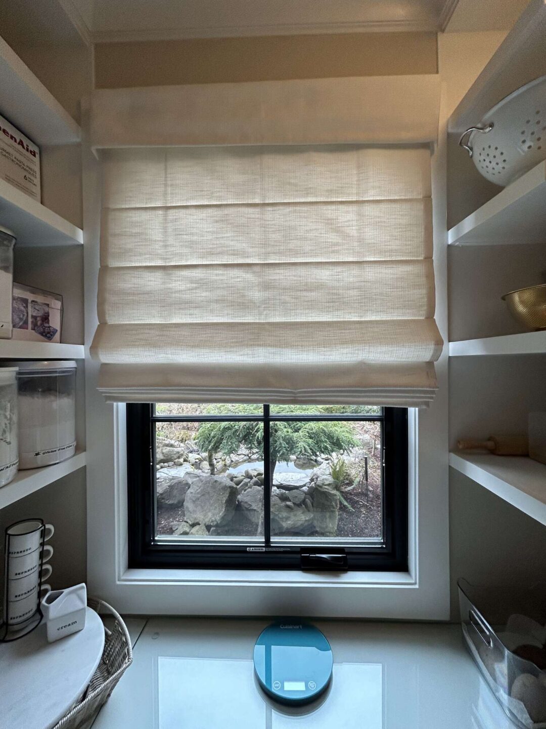 A small window with a beige Roman shade is centered between shelves holding kitchen items in this Portland kitchen. A digital kitchen scale sits on the counter. Pearl Painters, local Portland painters, can enhance spaces like these with professional painting services.