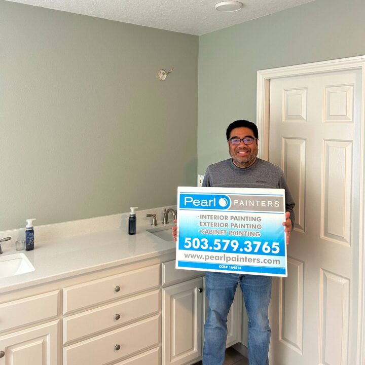Man holds Pearl Painters sign with contact info in a bathroom with white cabinets, sink, and light green wall. Painter in Portland.