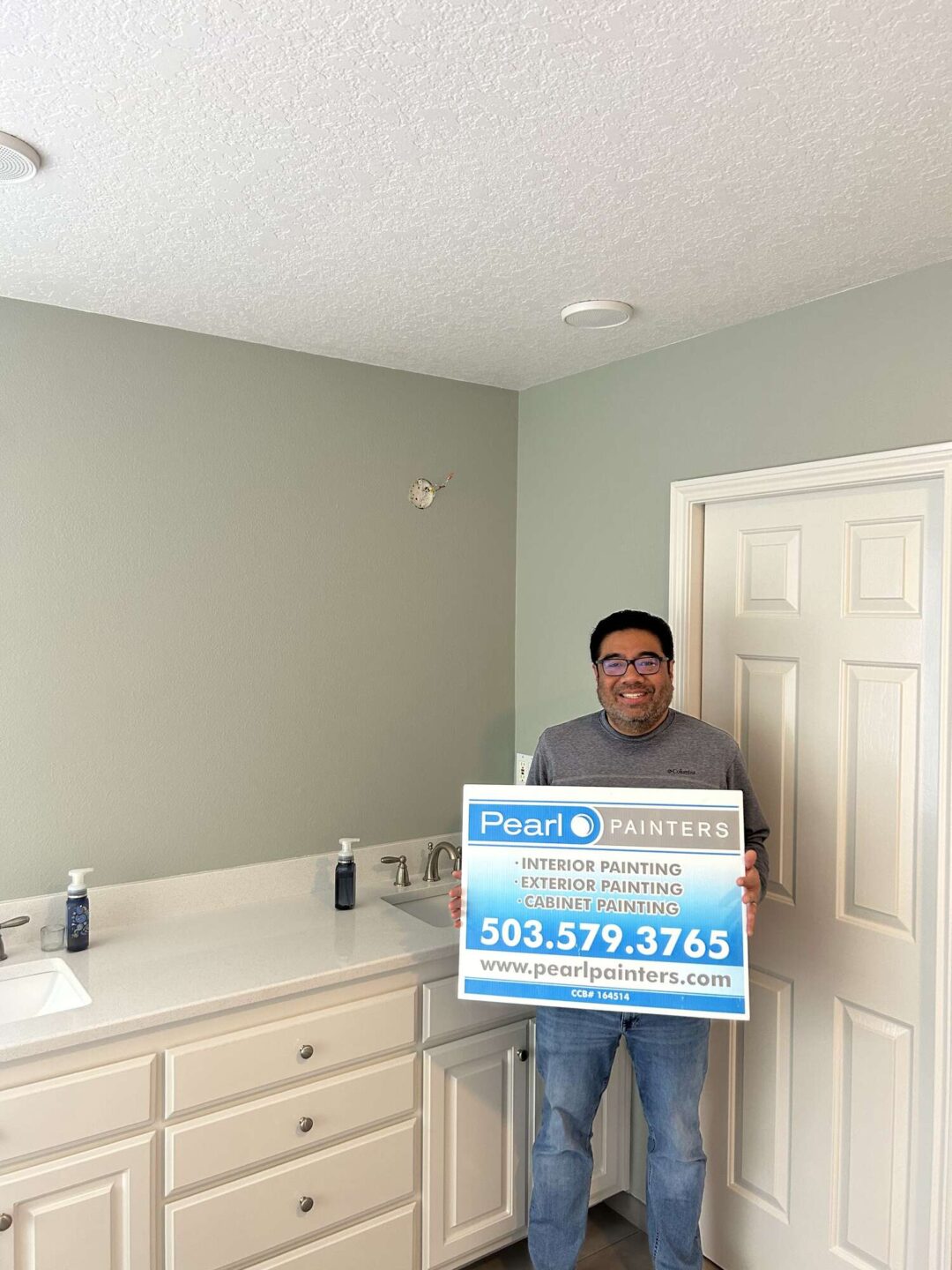 A painter stands in a light-colored bathroom with a double sink and white door, holding a Pearl Painters sign with contact info. Photo by Mallen, Portland, April 2025.