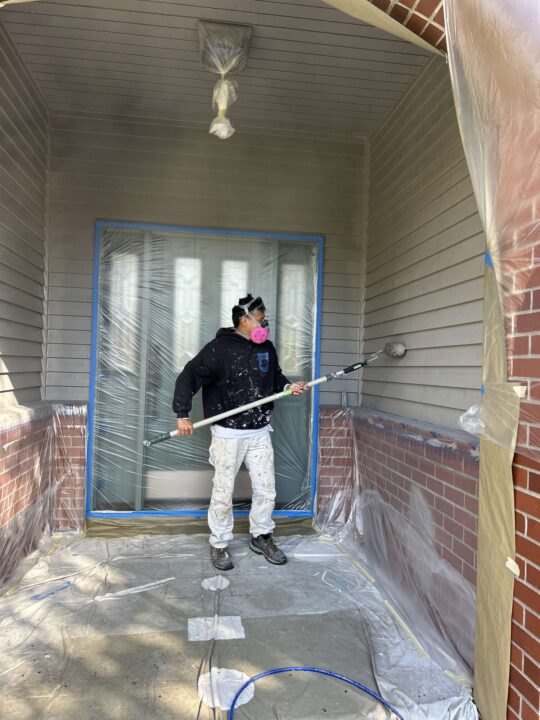 In April 2025, painter Scott from Pearl Painters in Portland spray paints a house exterior, wearing protective gear and using plastic sheets to cover the area.