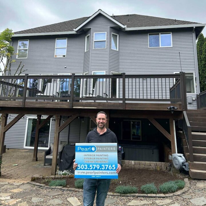Man stands by gray two-story house in Portland, holding Pearl Painters sign for interior and exterior painting services.
