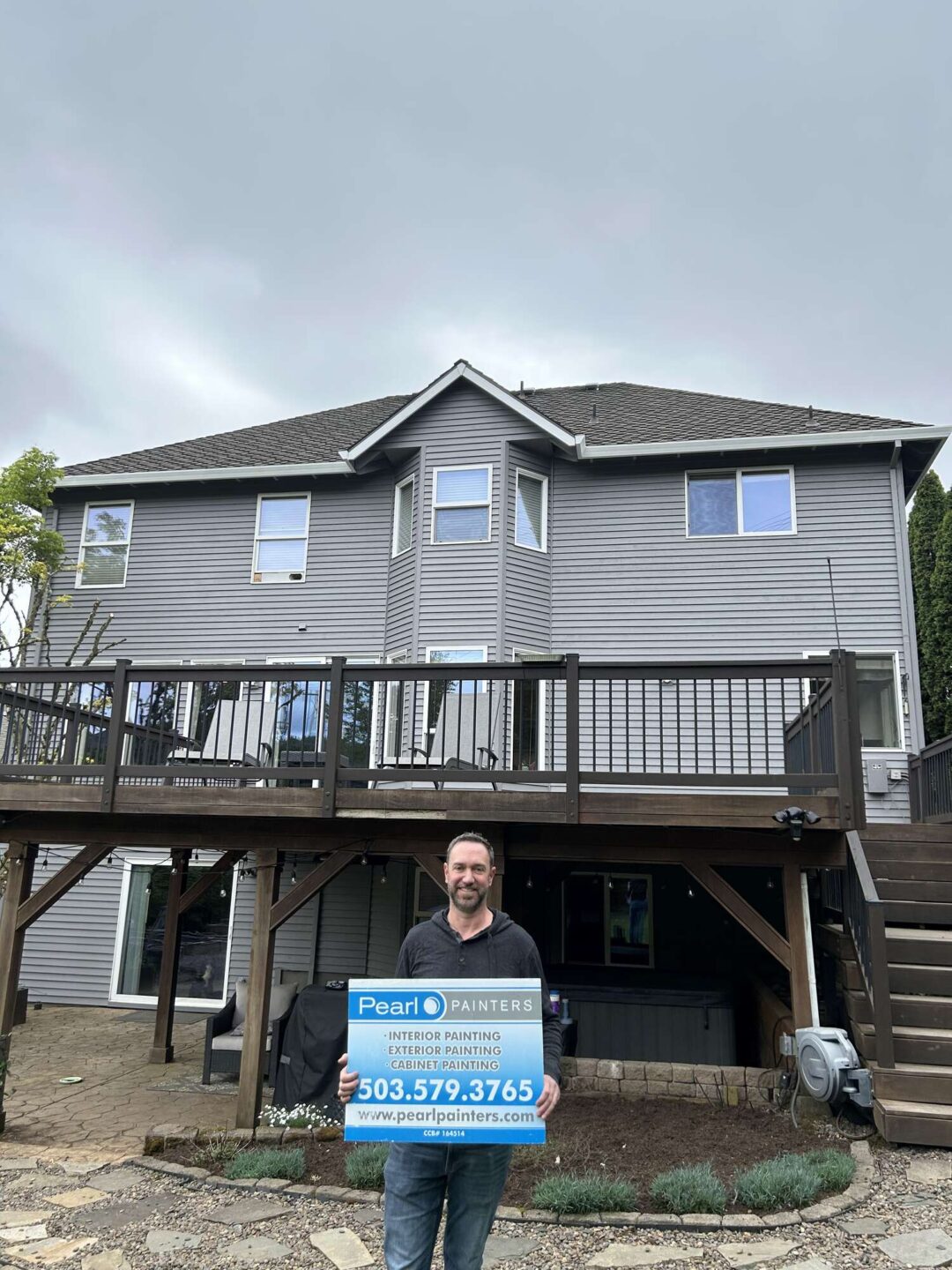Scott holds a Pearl Painters sign in front of a two-story gray house with a wooden deck in Portland, advertising interior and exterior painting for April 2025.