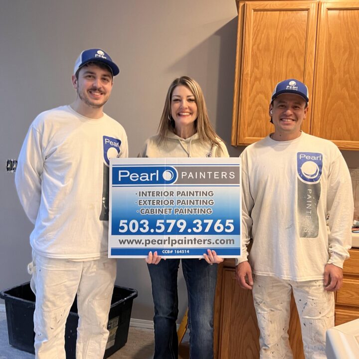 Three people, two in painter uniforms, hold a Pearl Painters painting service sign with contact info indoors in Portland.
