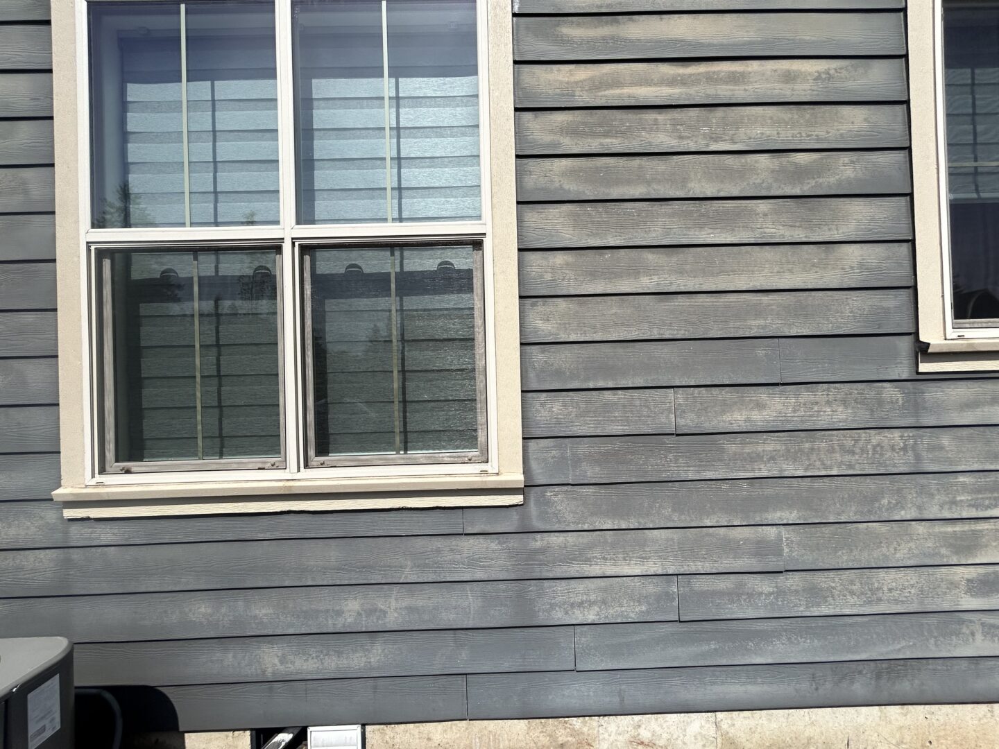 A gray wooden house exterior with two windows, one partially in the frame on the right side. Shadows and reflections are visible on the glass. A small section of an air conditioning unit is visible, highlighting subtle exterior paint failures in the background.