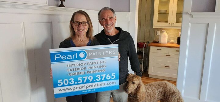 Two people stand in a kitchen holding a Pearl Painters (Portland) sign with contact details; a brown poodle is in front of them.