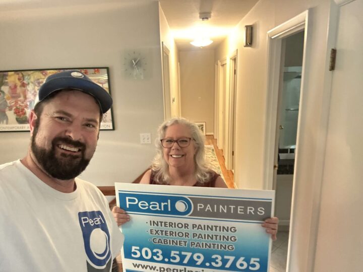 Two people stand indoors in Portland, smiling at the camera. One holds a Pearl Painters sign advertising interior, exterior, and cabinet painting services with contact information.