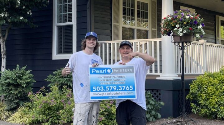 Two painters from Pearl Painters stand outside a Portland house holding a sign with their painter services and contact info; a porch and flowers are in the background.