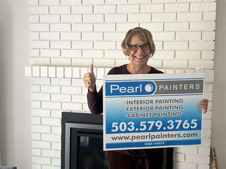 A woman stands in front of a white brick fireplace, smiling and giving a thumbs up while holding a Pearl Painters sign displaying painting services and contact information in Portland.