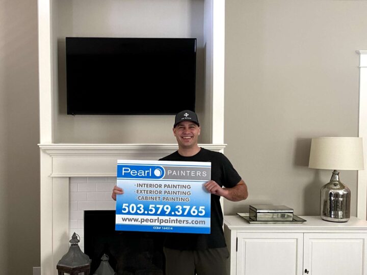 A painter in Portland stands in front of a fireplace holding a blue Pearl Painters sign with contact details and services.