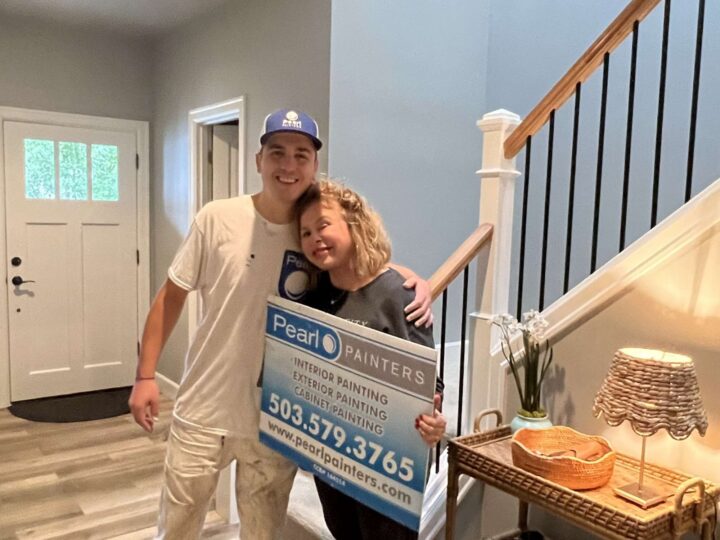 Two people stand in a home entryway in Portland, holding a Pearl Painters sign for interior and exterior painting services.