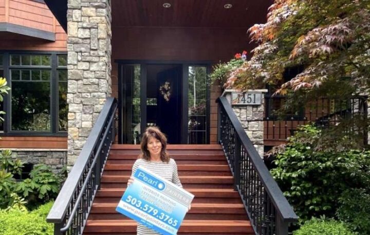 A woman stands on the front steps of a stone and wood house in Portland, holding a Pearl Painters Certification sign with a phone number.