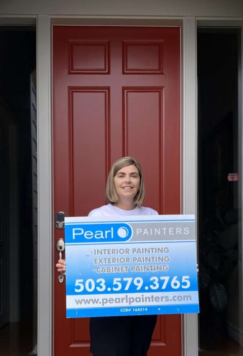 A woman stands in front of a red door in Portland holding a Pearl Painters sign advertising painting services.