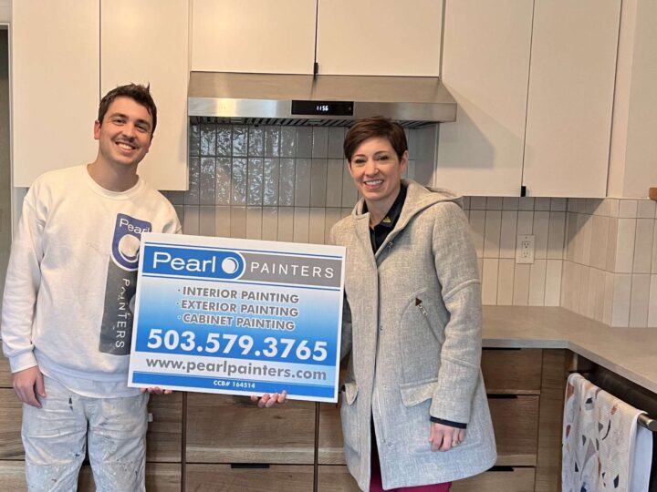 Two people in a kitchen in Portland hold a Pearl Painters sign advertising interior, exterior, and cabinet painting services. The painter’s contact info and website are displayed.