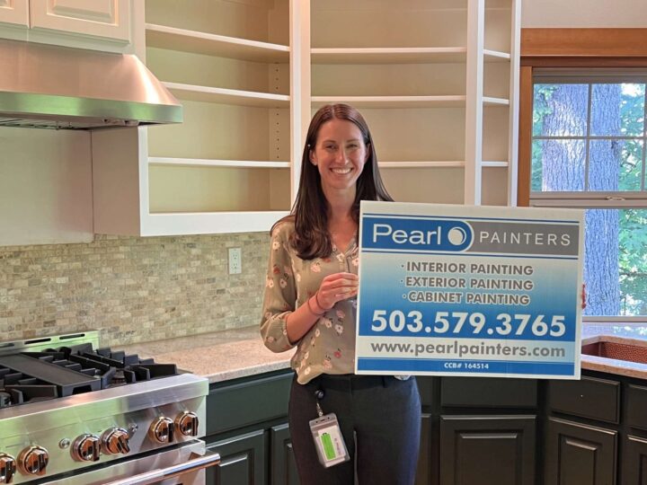 A woman in a kitchen in Portland holds a Pearl Painters sign advertising painting services; empty shelves and a stove are visible.