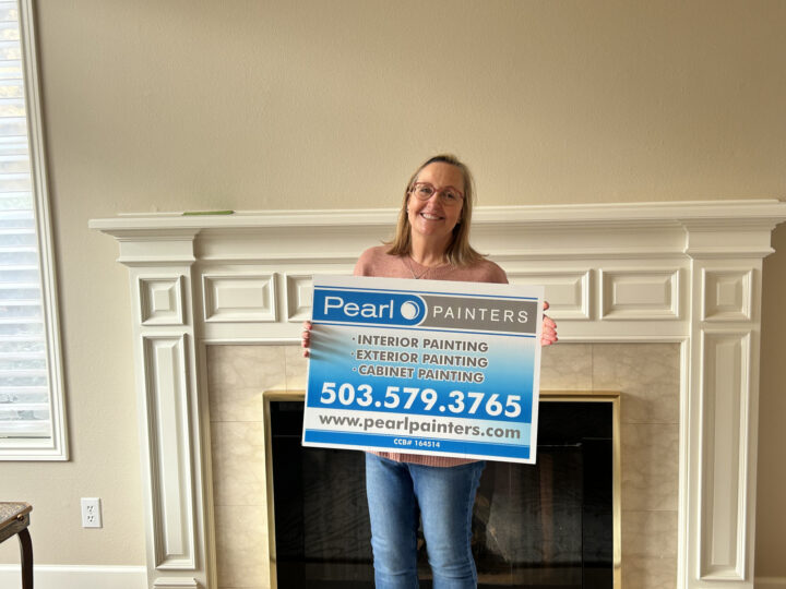 A woman stands indoors in Portland, holding a Pearl Painters sign advertising painting services in front of a white fireplace.